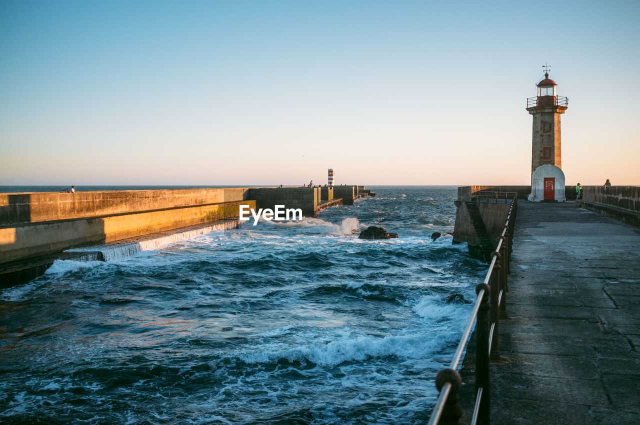 Footpath leading towards lighthouse in sea against sky during sunset