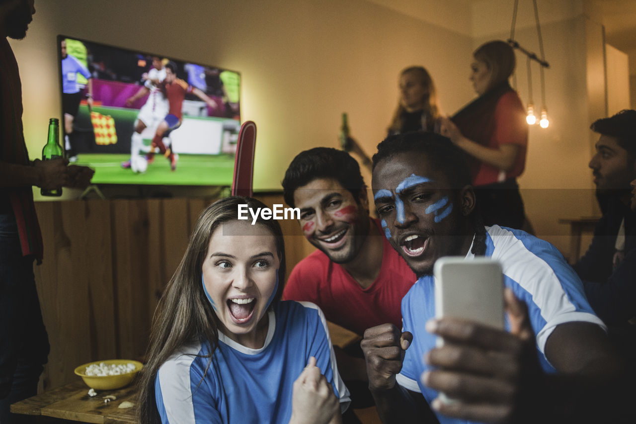 Fans taking selfie with mobile phone while friends watching soccer match on tv