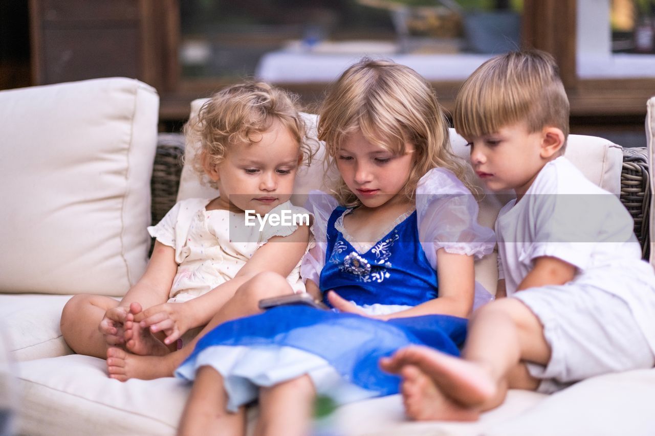 Girl with siblings using phone while sitting on sofa at home