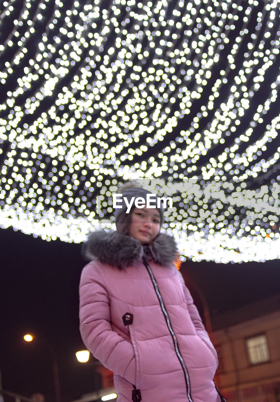 one person, adult, clothing, women, warm clothing, winter, standing, lifestyles, leisure activity, front view, architecture, low angle view, smiling, waist up, portrait, happiness, three quarter length, young adult, outdoors, cold temperature, looking, illuminated, built structure, night, nature, music, emotion, technology, looking at camera, spring, arts culture and entertainment, building exterior, city, casual clothing