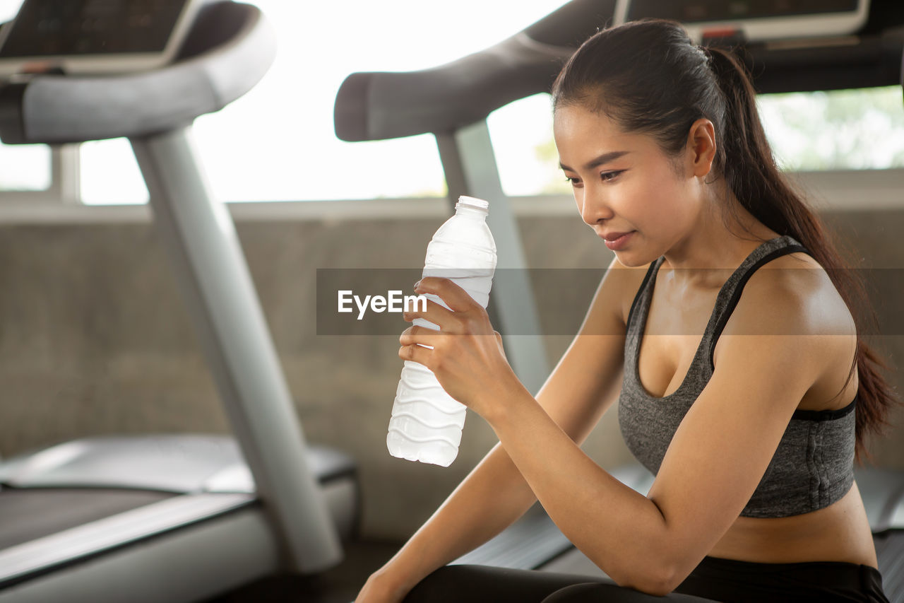 Woman holding water bottle while sitting on treadmill in gym