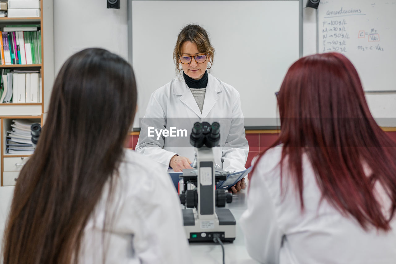 Adult female in white robe reading notes near microscope while teaching young women in modern laboratory in university