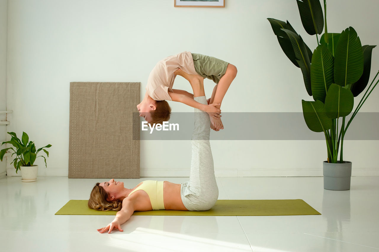 A woman, doing yoga with a child boy, lies on a sports mat, raising her legs up