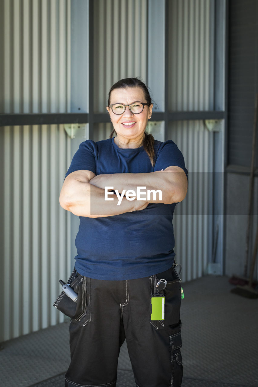 Portrait of smiling mature female worker standing with arms crossed on warehouse entrance