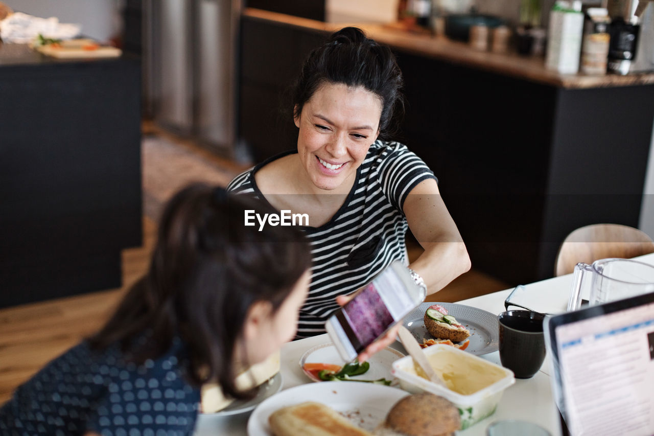 Smiling mother showing mobile phone to daughter while having breakfast at dining table