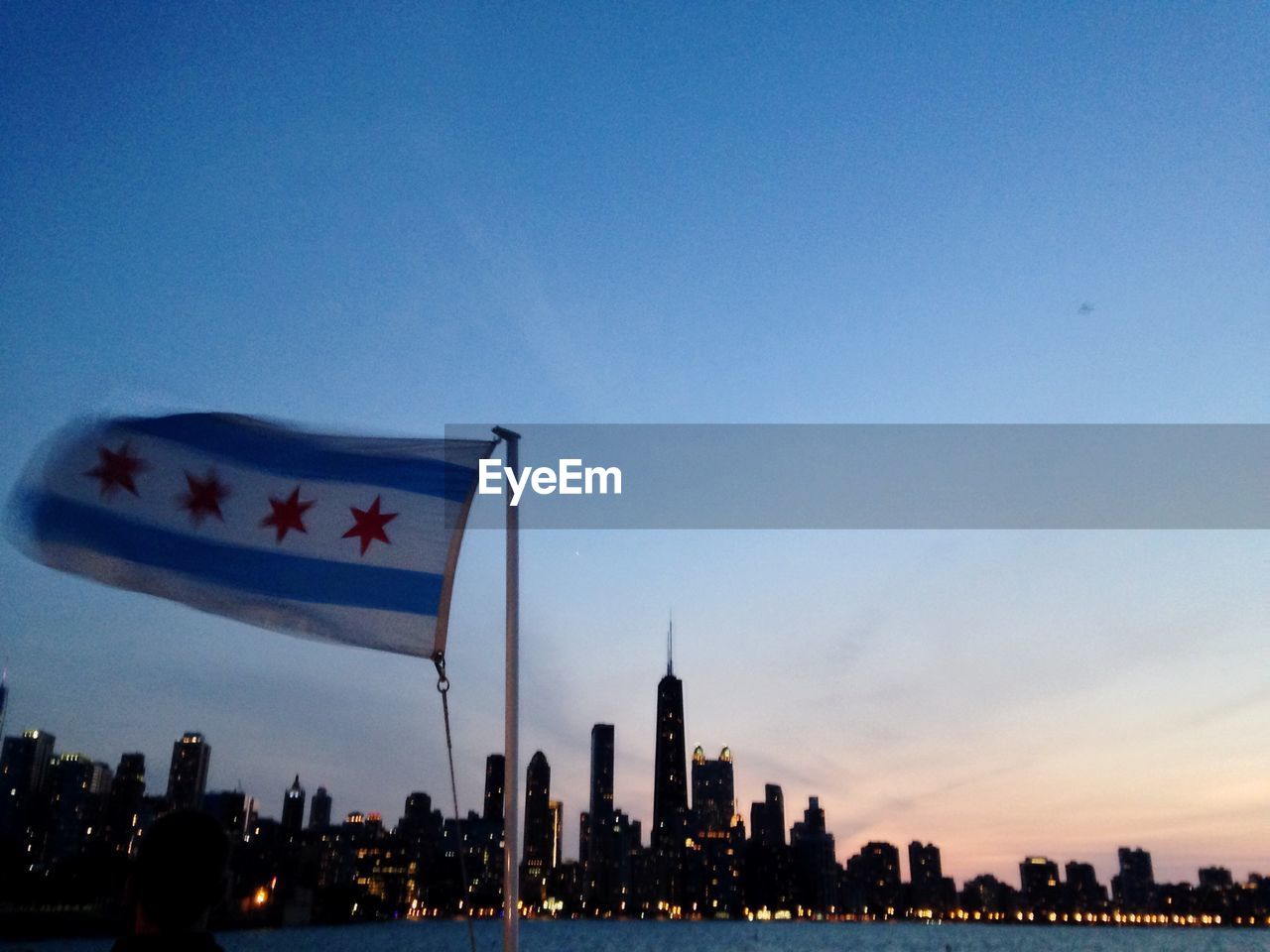 Flag of chicago waving by river against city during sunset