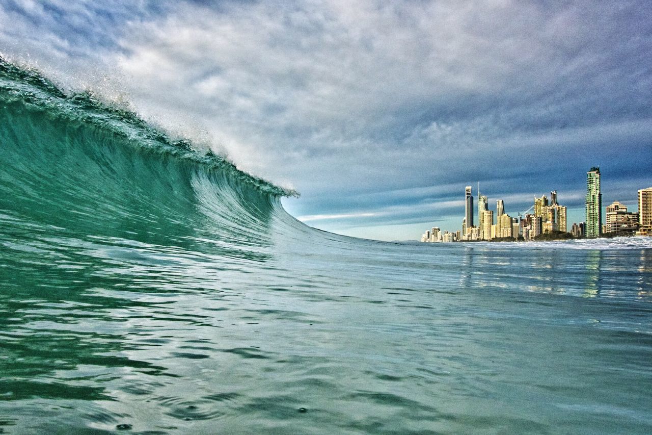 Idyllic view of sea wave by city against cloudy sky