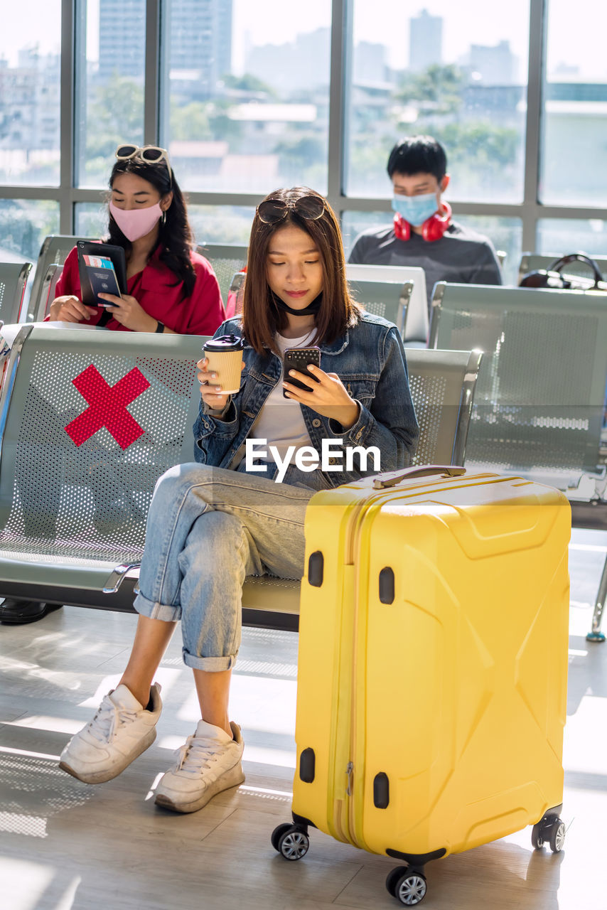 Asian young tourist girl using phone and hold hot coffee glass at departure waiting seats at airport