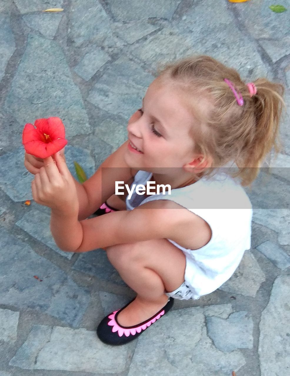 High angle view of cute girl holding red flower while crouching on footpath