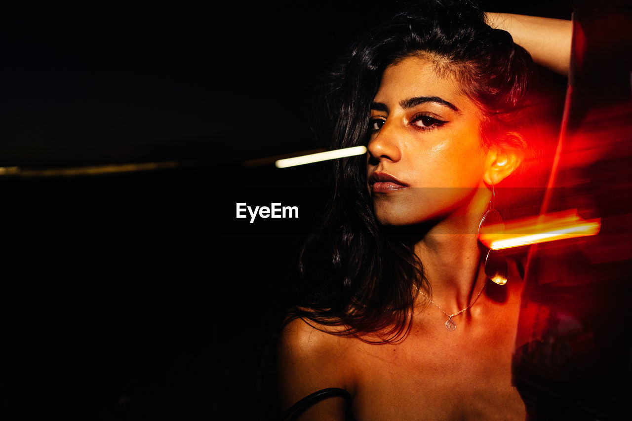 Confident young hispanic female with long wavy hair and bare shoulders keeping hand on head and looking at camera while standing near glass window with light reflections in darkness at night time