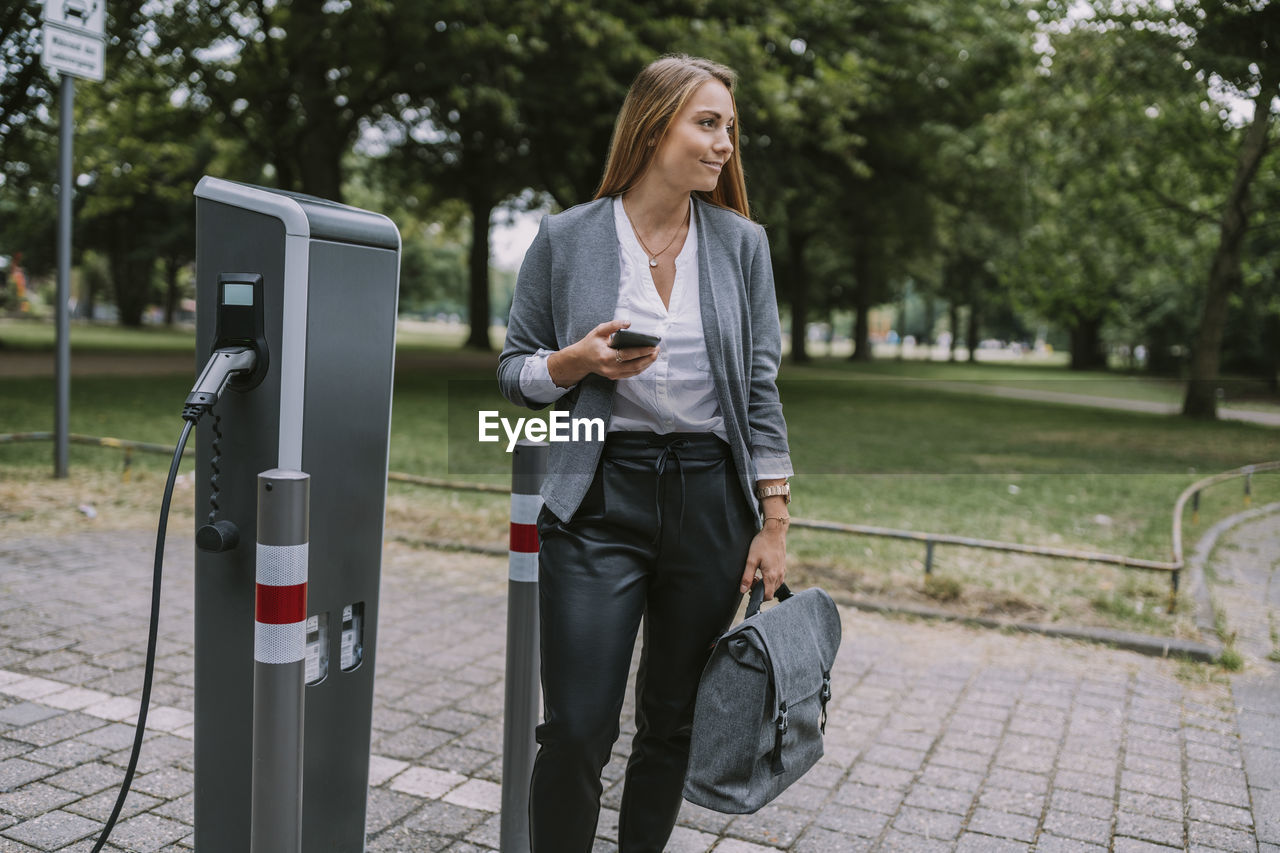 Businesswoman with mobile phone standing by electric charging station