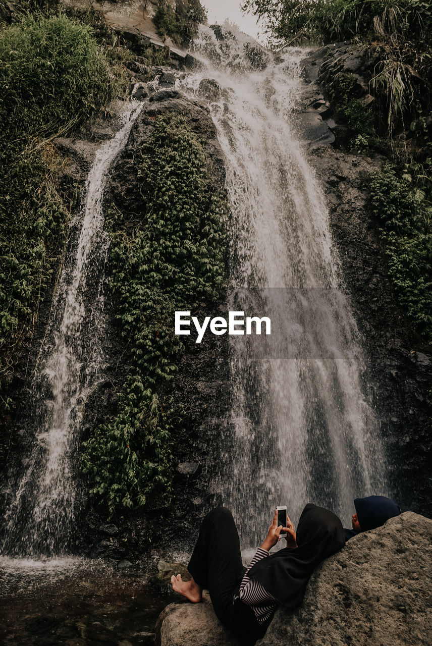 Couple lying down on rock while using mobile phone against waterfall