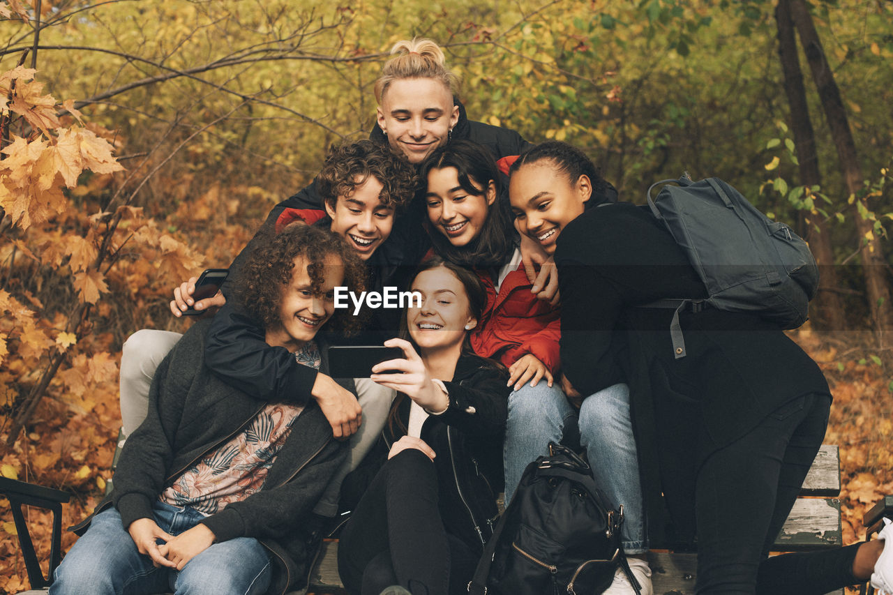 Teenage girl taking selfie with friends while sitting on bench against trees during autumn