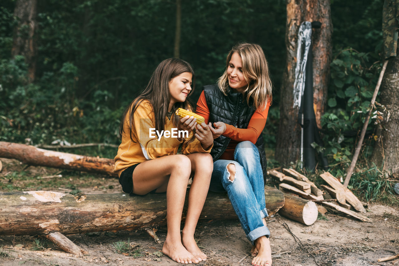 A teenage girl with her mother is sitting on a log in the forest, a girl is eating corn
