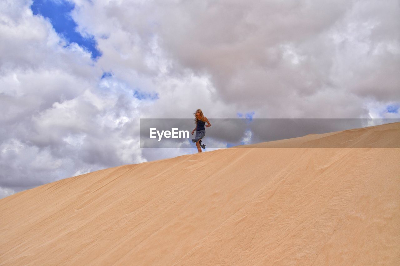 Low angle view of woman walking on sand dune against cloudy sky
