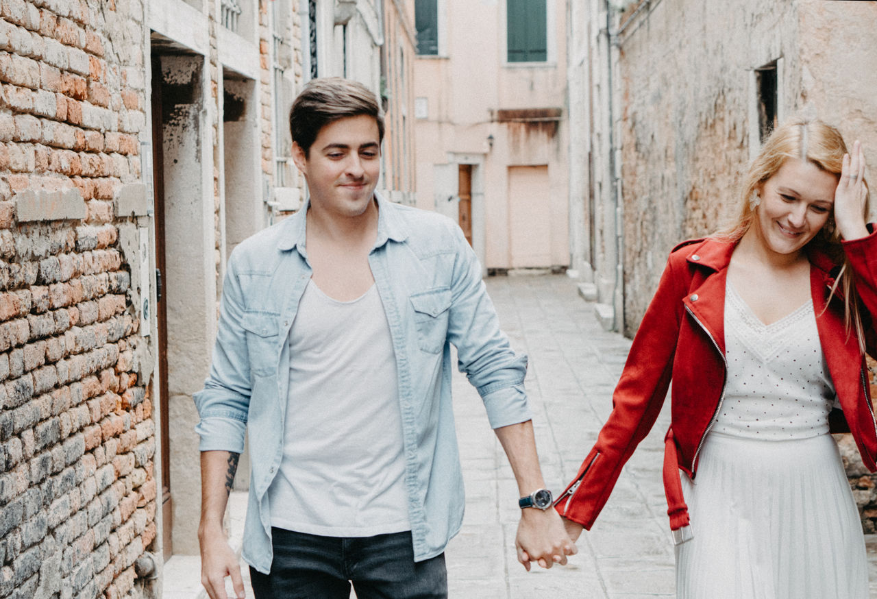 Smiling young couple holding hands while walking on footpath against buildings in city