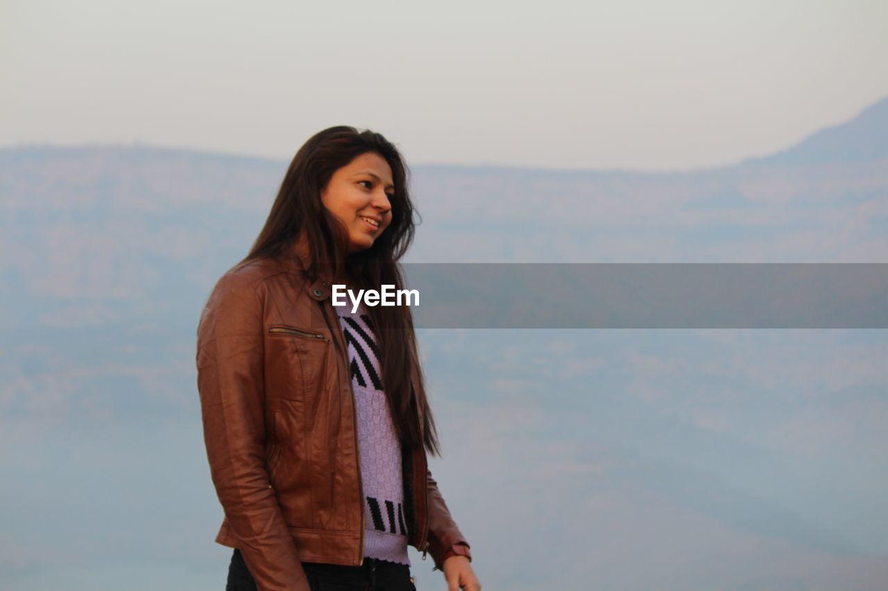 Smiling young woman wearing jacket standing against mountain