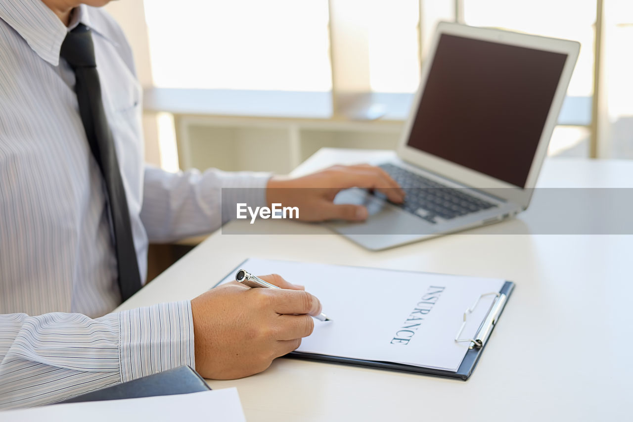 Midsection of insurance agent writing on paper while using laptop on table at office