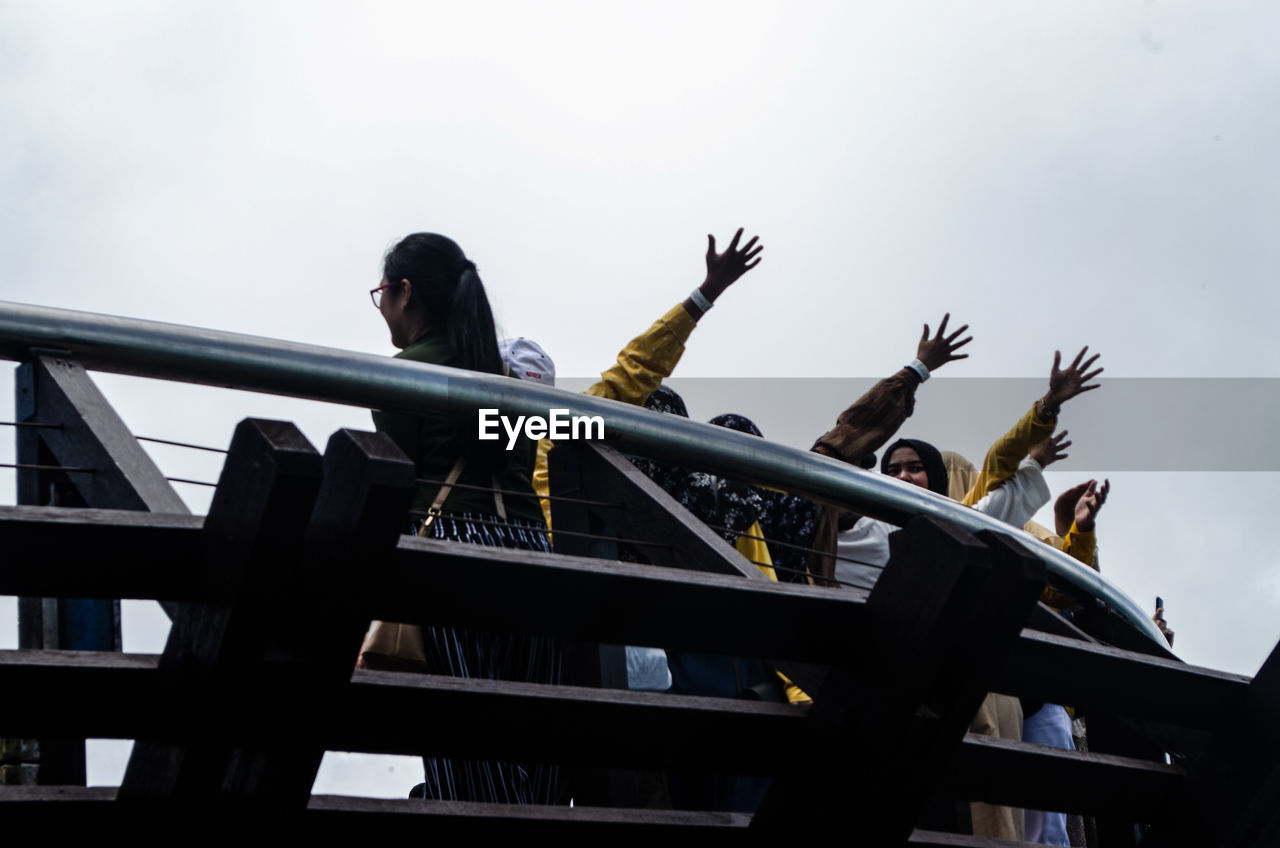 Low angle view of women with arms raised by railing against sky