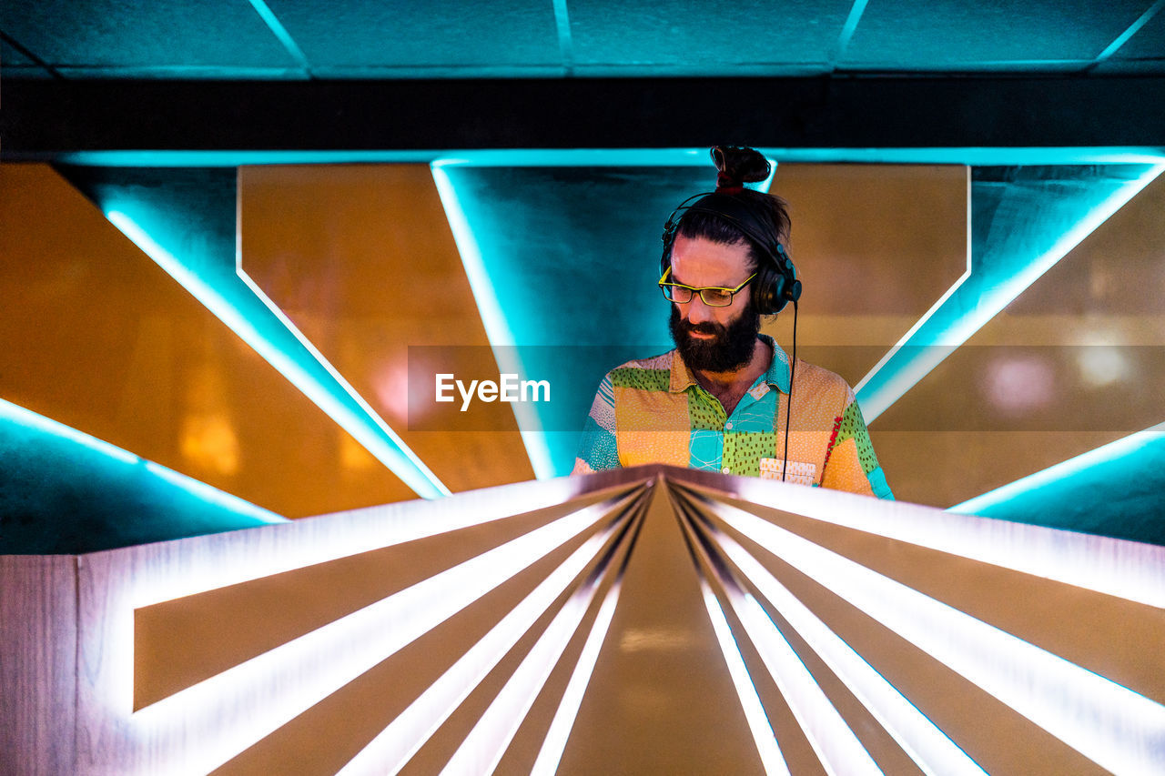 Concentrated young bearded male dj in eyeglasses and headphones standing at illuminated counter in modern nightclub