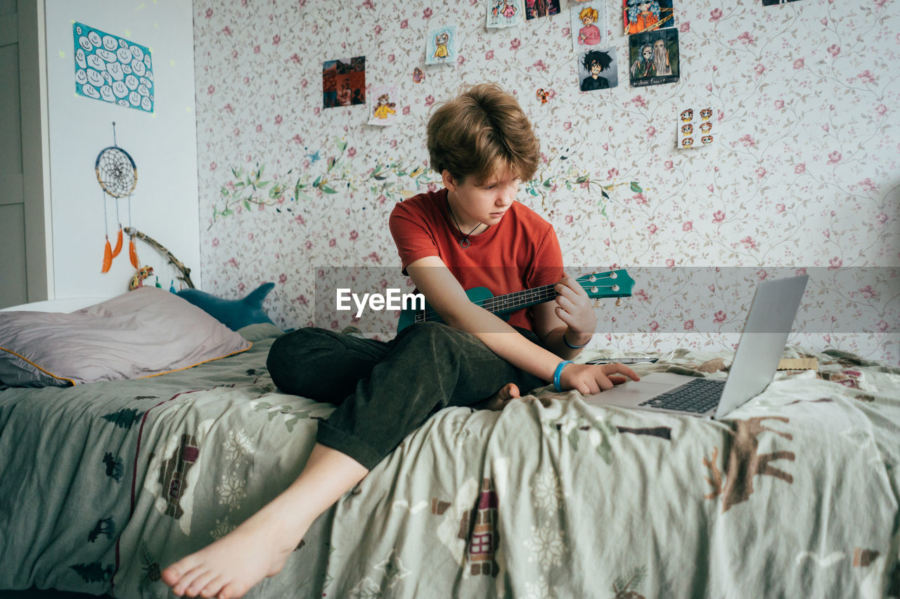 Teenage girl sitting on bed learning to play ukulele using online lessons on laptop.