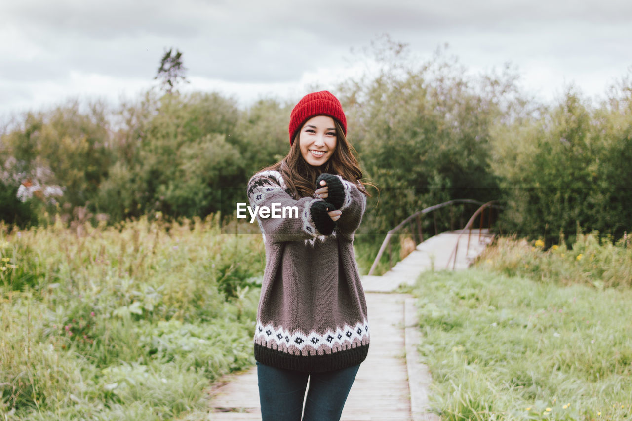 Portrait of smiling young woman standing on footpath against trees and plants