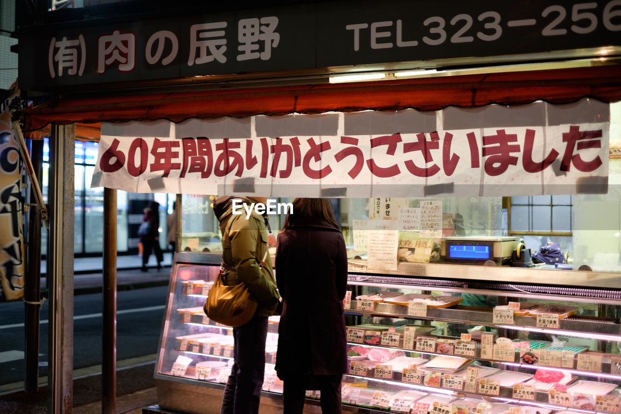 Midsection of friends standing near illuminated meat shop in city at night