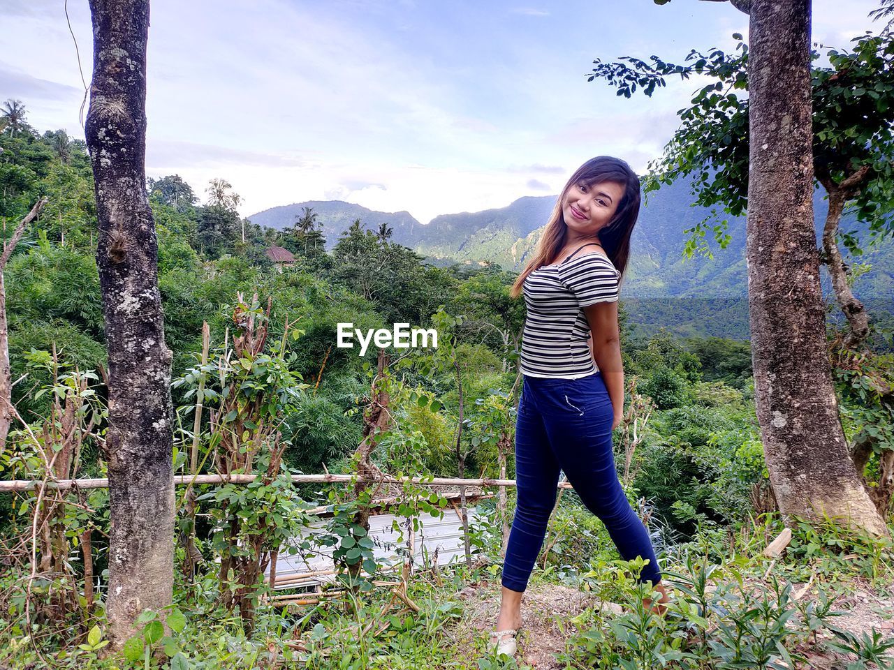 FULL LENGTH PORTRAIT OF YOUNG WOMAN STANDING ON TREE AGAINST SKY