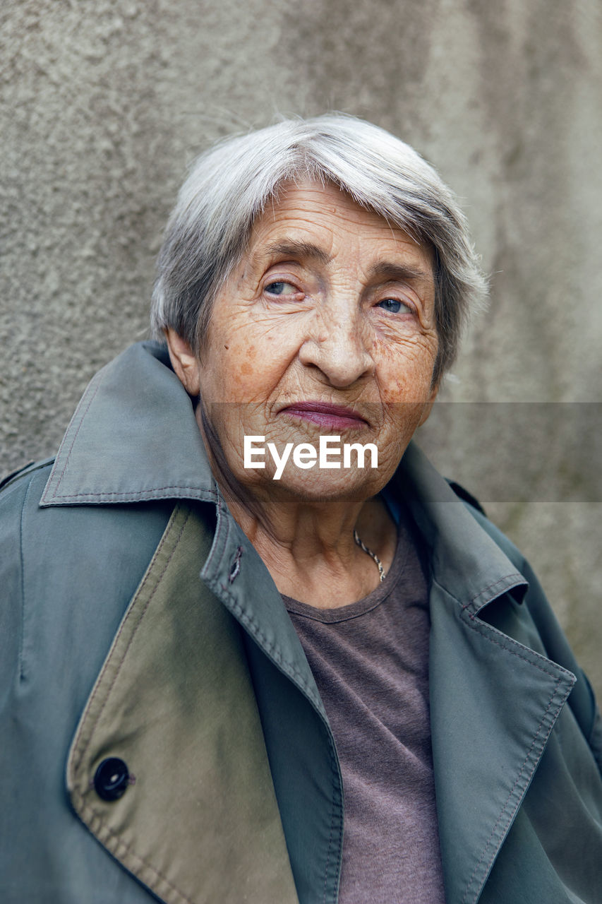 Portrait of an old siniors grandmother of 90 with gray hair stands against a gray wall on the street