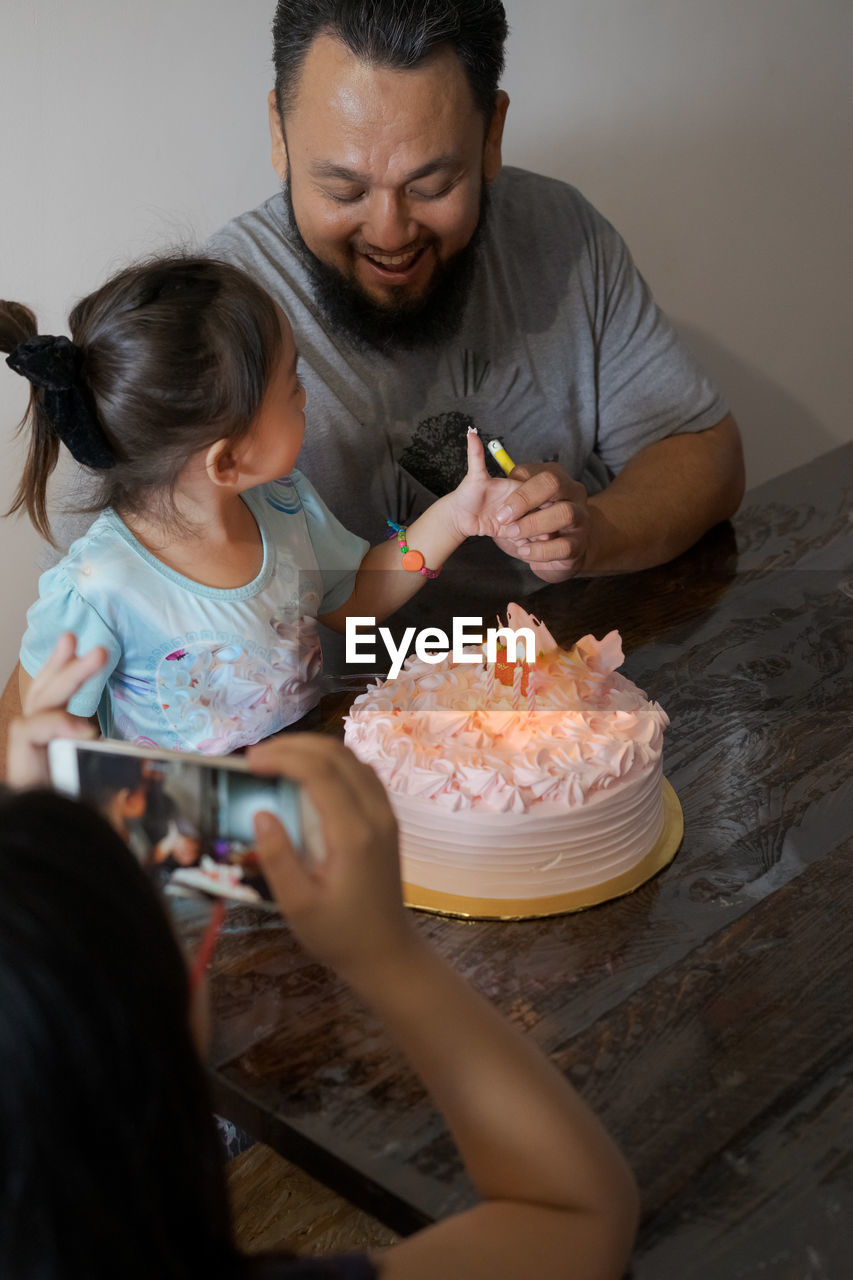Girl photographing father and daughter with birthday cake on table