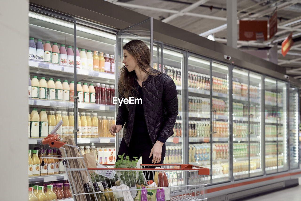 Woman standing with shopping cart while looking at bottles in refrigerated cabinet