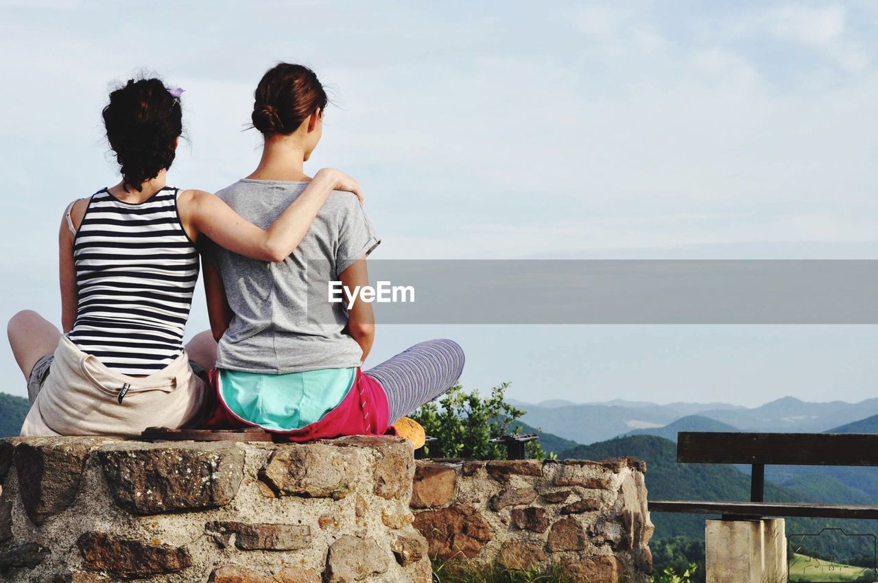 Lesbian couple sitting on wall against sky