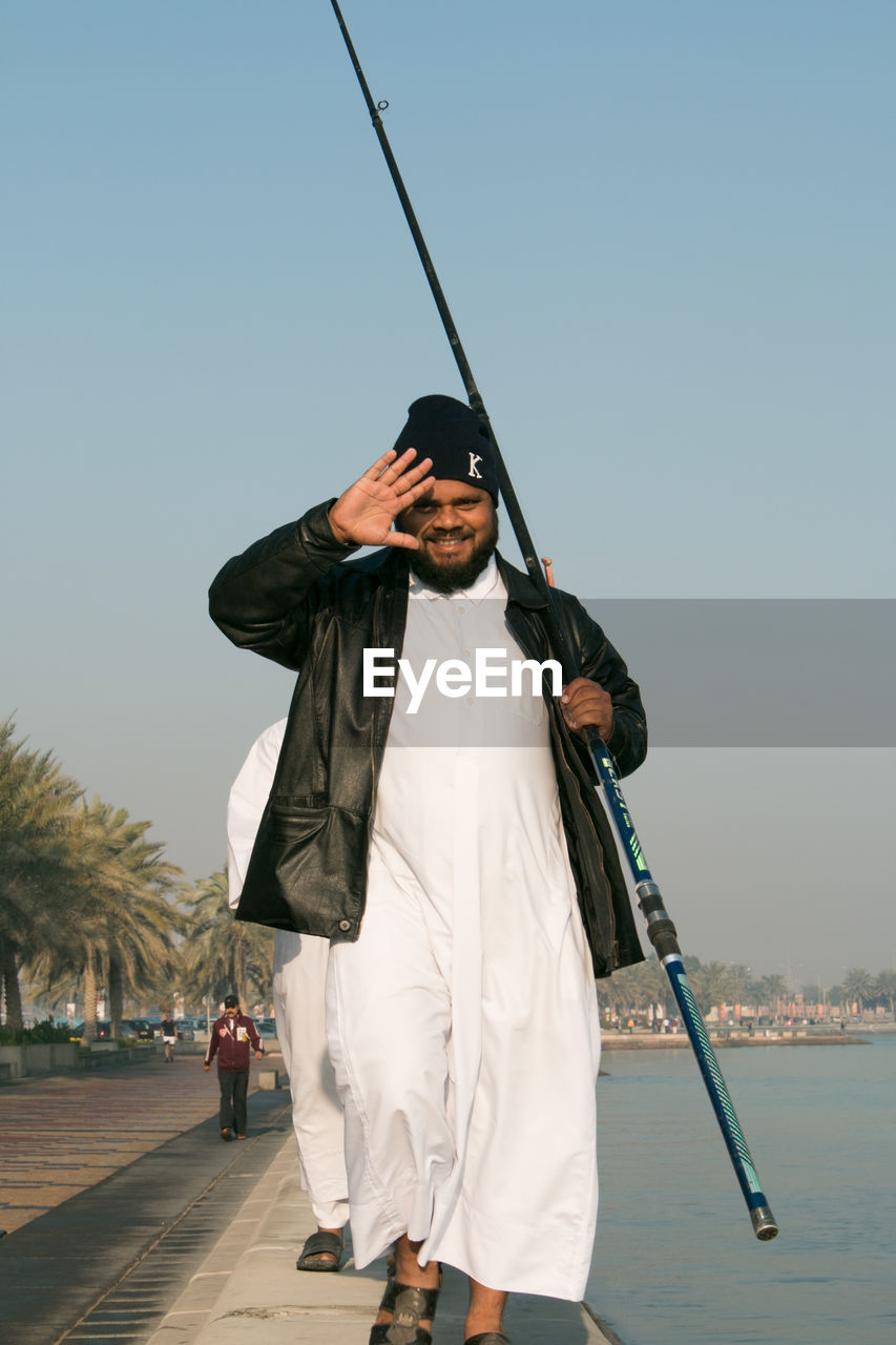 Portrait of happy man with fishing rod walking on promenade against sky