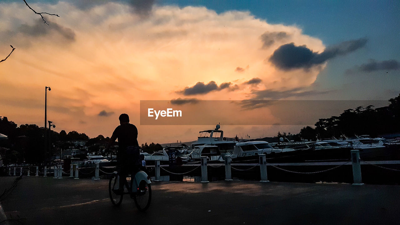 Rear view of silhouette man riding bicycle on street against sky during sunset