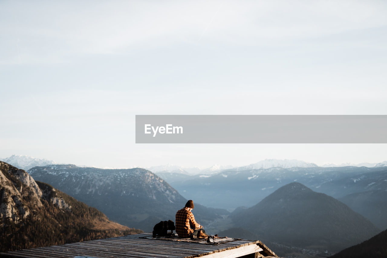 Back view of unrecognizable male tourist sitting on blanket at viewpoint and enjoying scenery of mountain ridge in morning