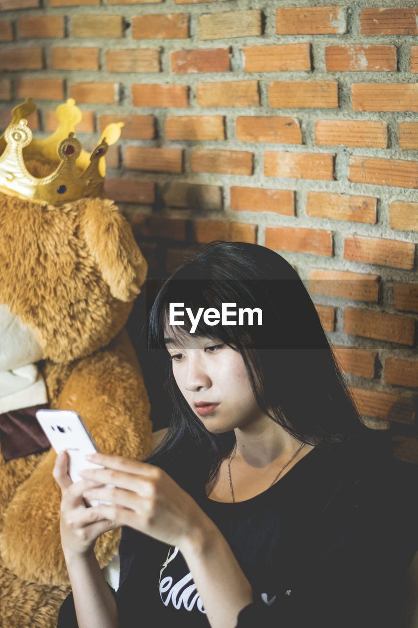 Young woman using phone while sitting by teddy bear on wall