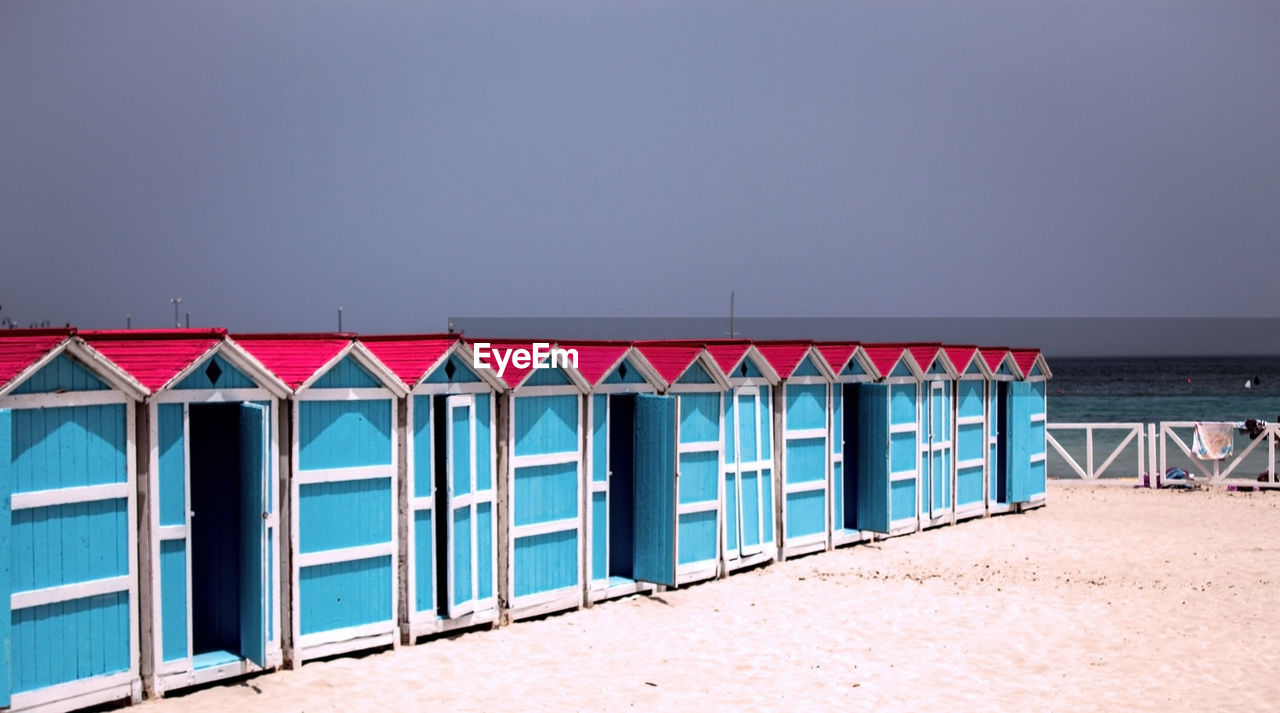 beach, land, sea, water, sand, sky, beach hut, nature, architecture, hut, in a row, no people, built structure, clear sky, horizon over water, multi colored, day, scenics - nature, horizon, tranquility, copy space, blue, outdoors, travel destinations, tranquil scene, beauty in nature, coastline, sunny, building exterior, protection, wood, holiday, side by side, sunlight