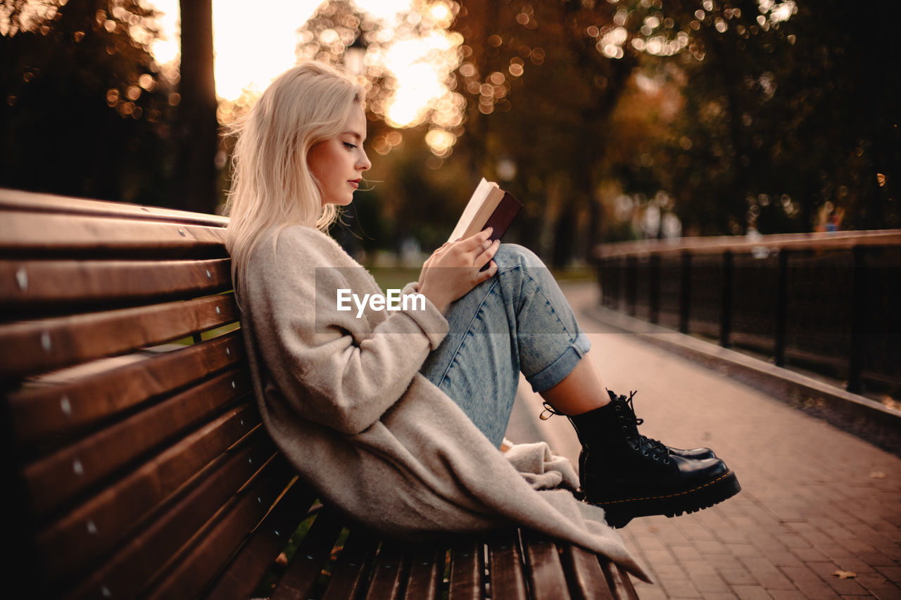 Teenage girl reading book sitting on bench in park during autumn