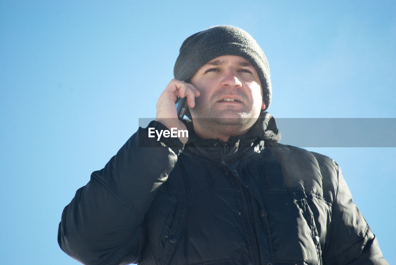 PORTRAIT OF HANDSOME YOUNG MAN USING SMART PHONE AGAINST SKY