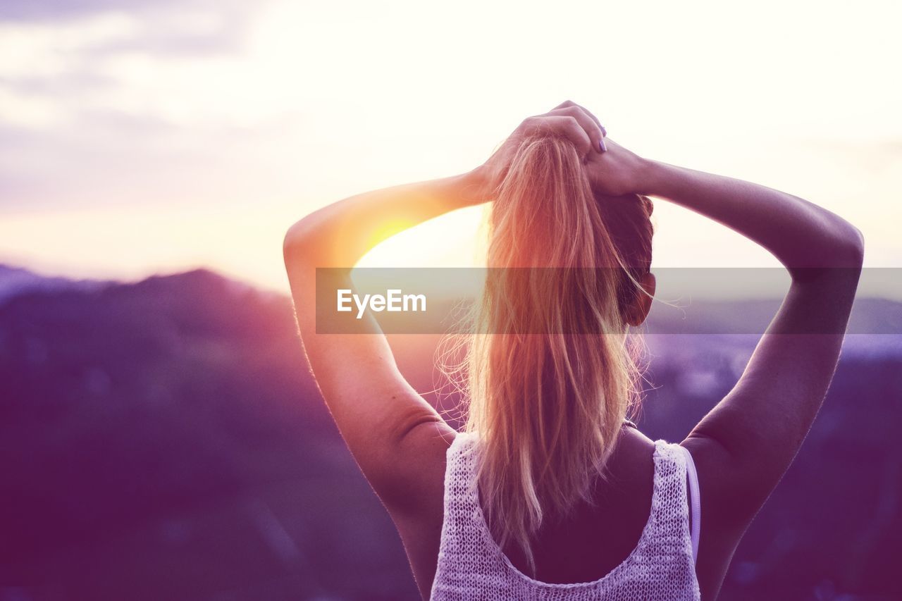 Rear view of woman tying ponytail against mountain during sunset