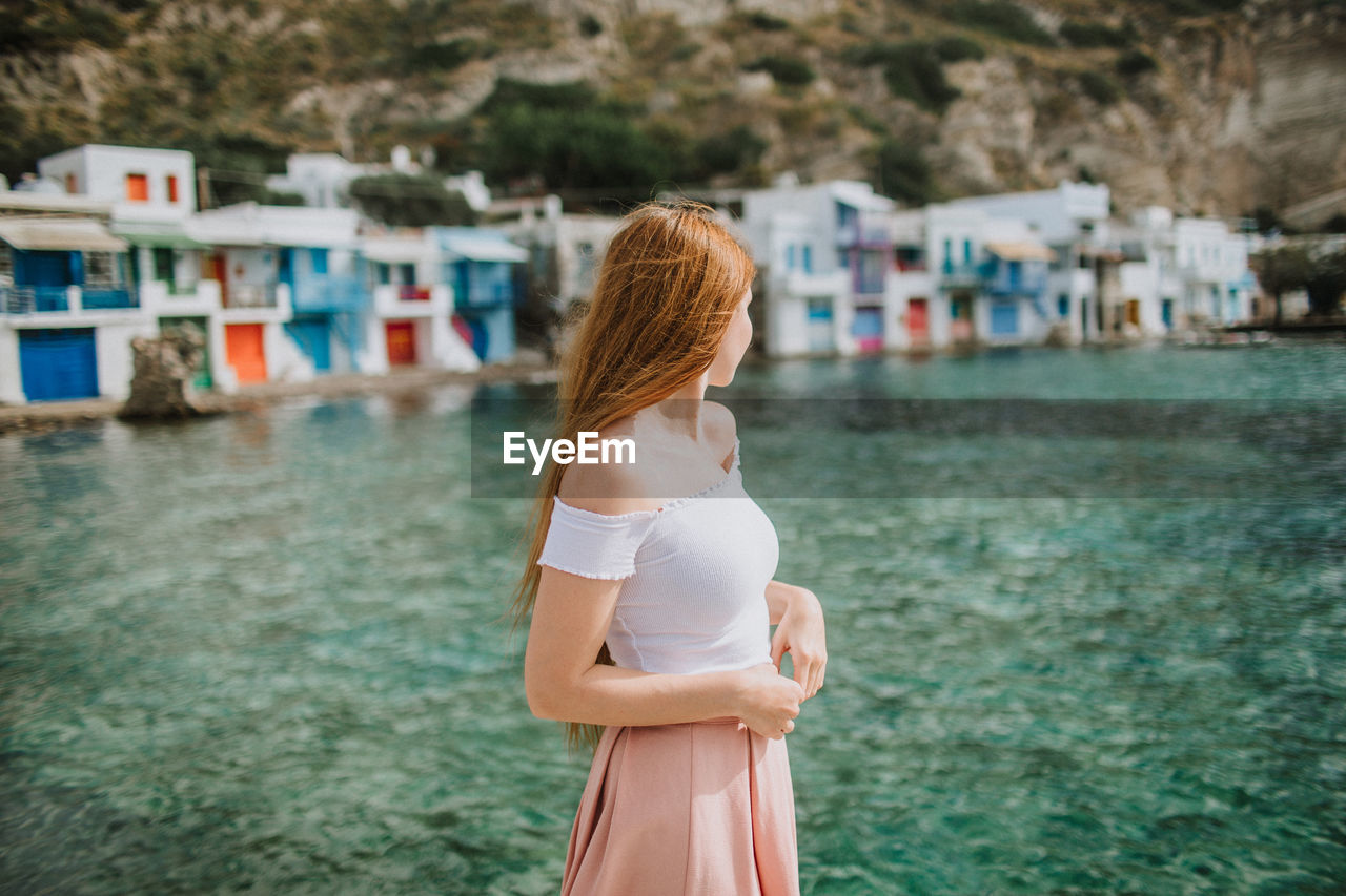 Side view anonymous female wearing stylish summer clothes standing in authentic coastal village with small low rise houses near crystal clear azure sea in sunny greece