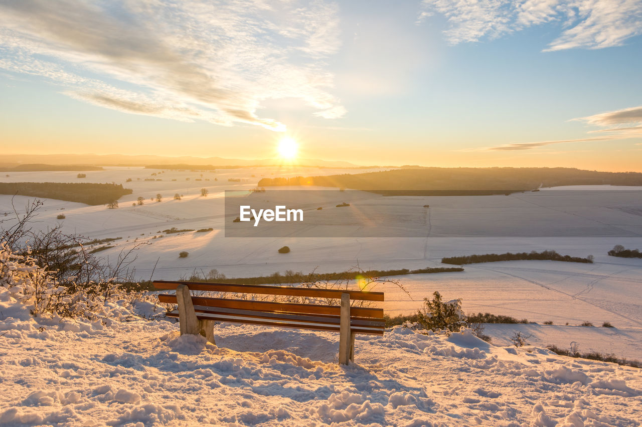 BENCHES ON SNOW COVERED FIELD AGAINST SKY DURING SUNSET