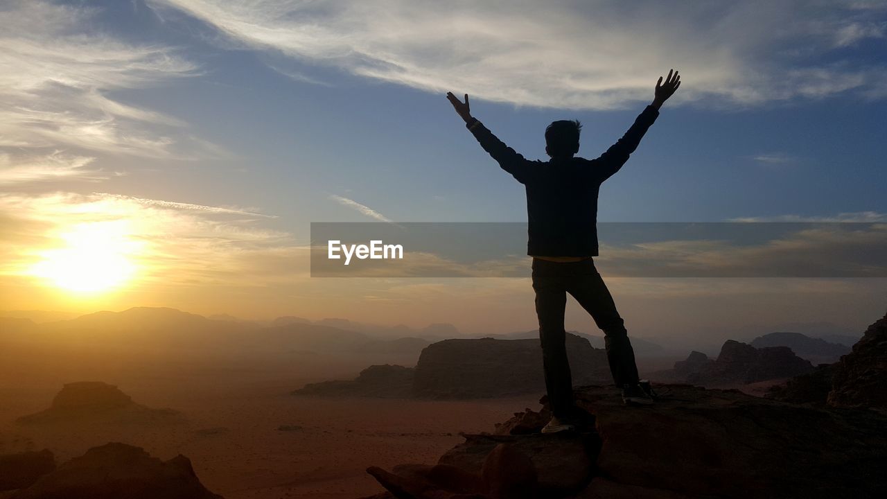 Boy standing on rock formation against sky during sunset