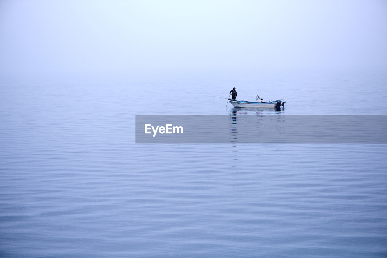 Man standing on moored fishing boat on calm lake during foggy weather