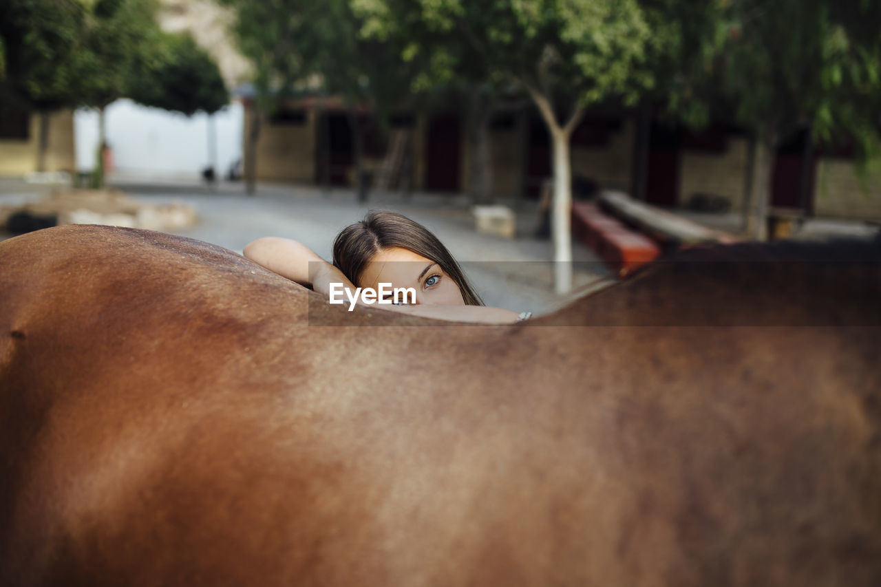 Portrait of young woman leaning on brown horse from behind