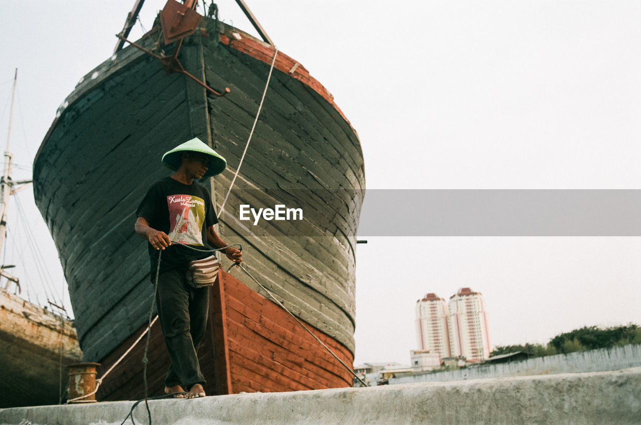 architecture, one person, adult, nature, sky, built structure, men, day, standing, hat, occupation, water, nautical vessel, transportation, outdoors, building exterior, full length, clothing, low angle view, vehicle, holding, ship, copy space, clear sky, working