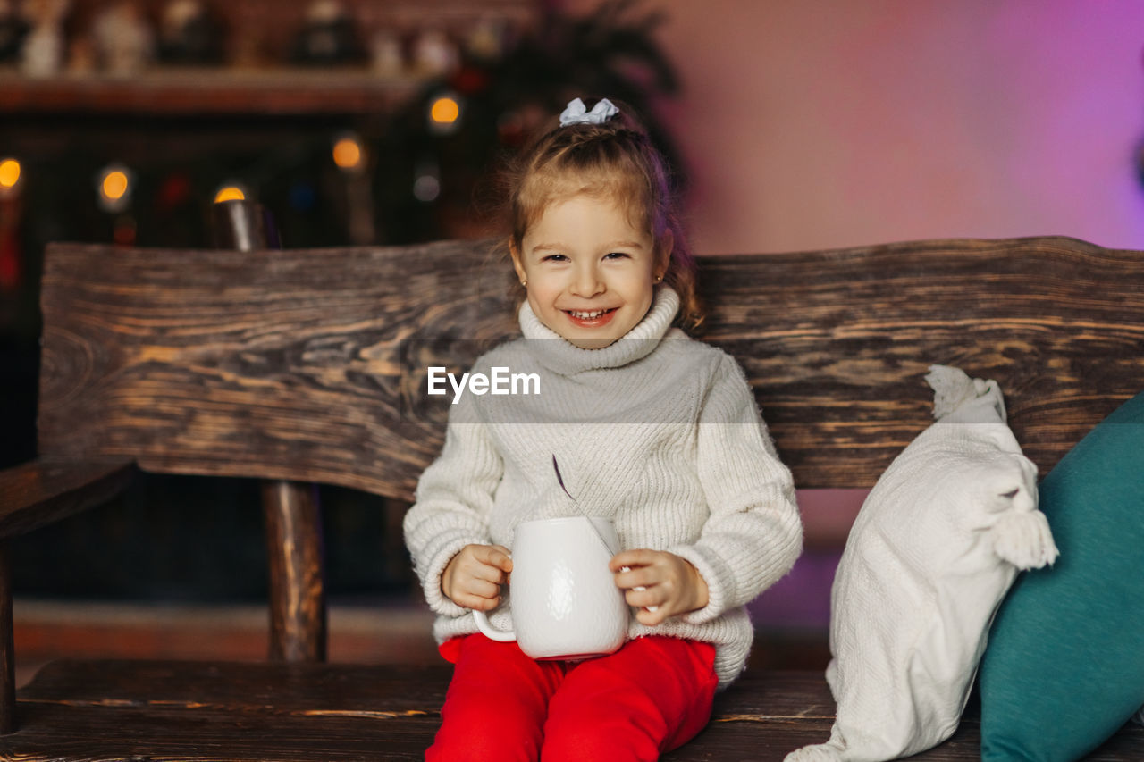 A cheerful baby is drinking hot chocolate on a bench by the fireplace. christmas mood