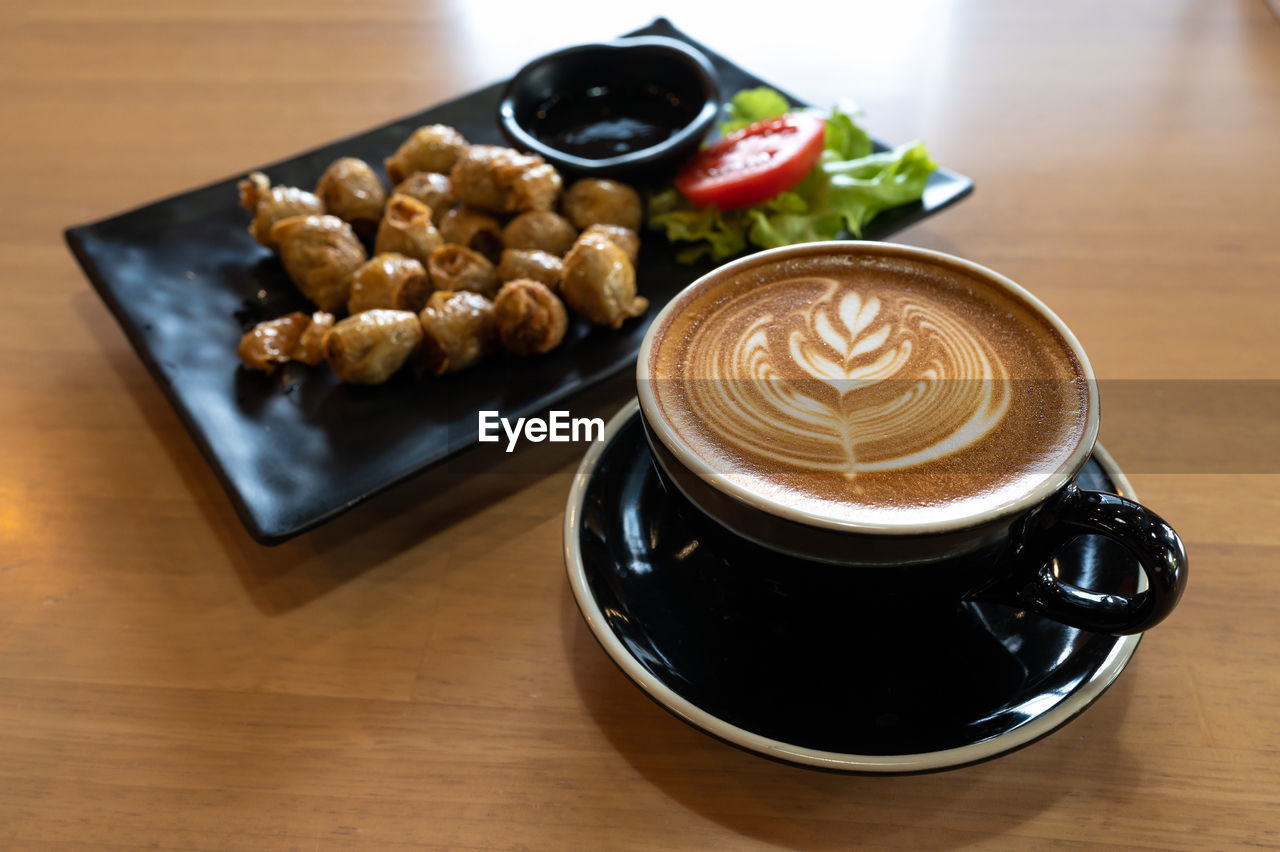 Latte art in black cup with snack on wooden table