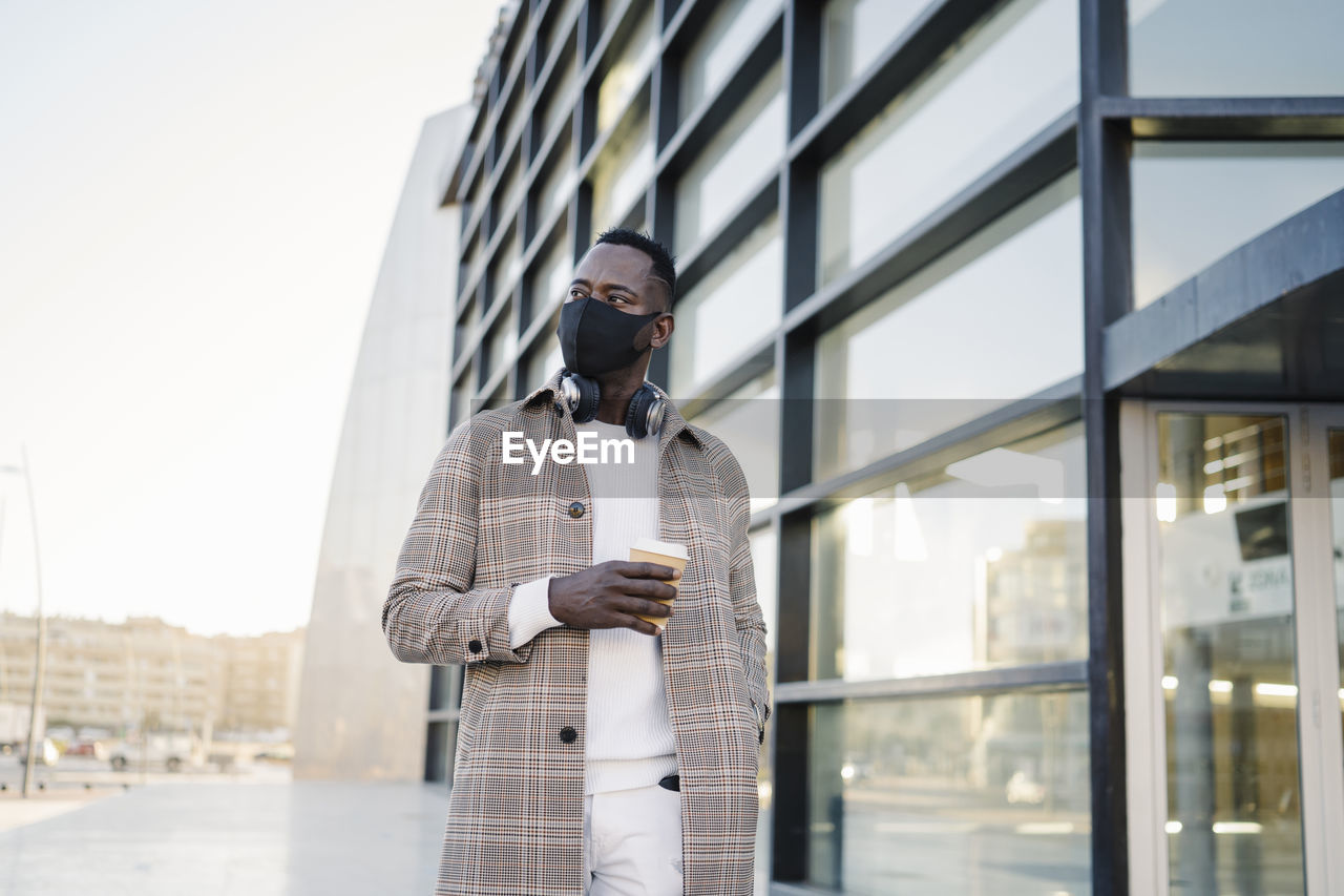 Fashionable man wearing protective face mask holding coffee cup against building