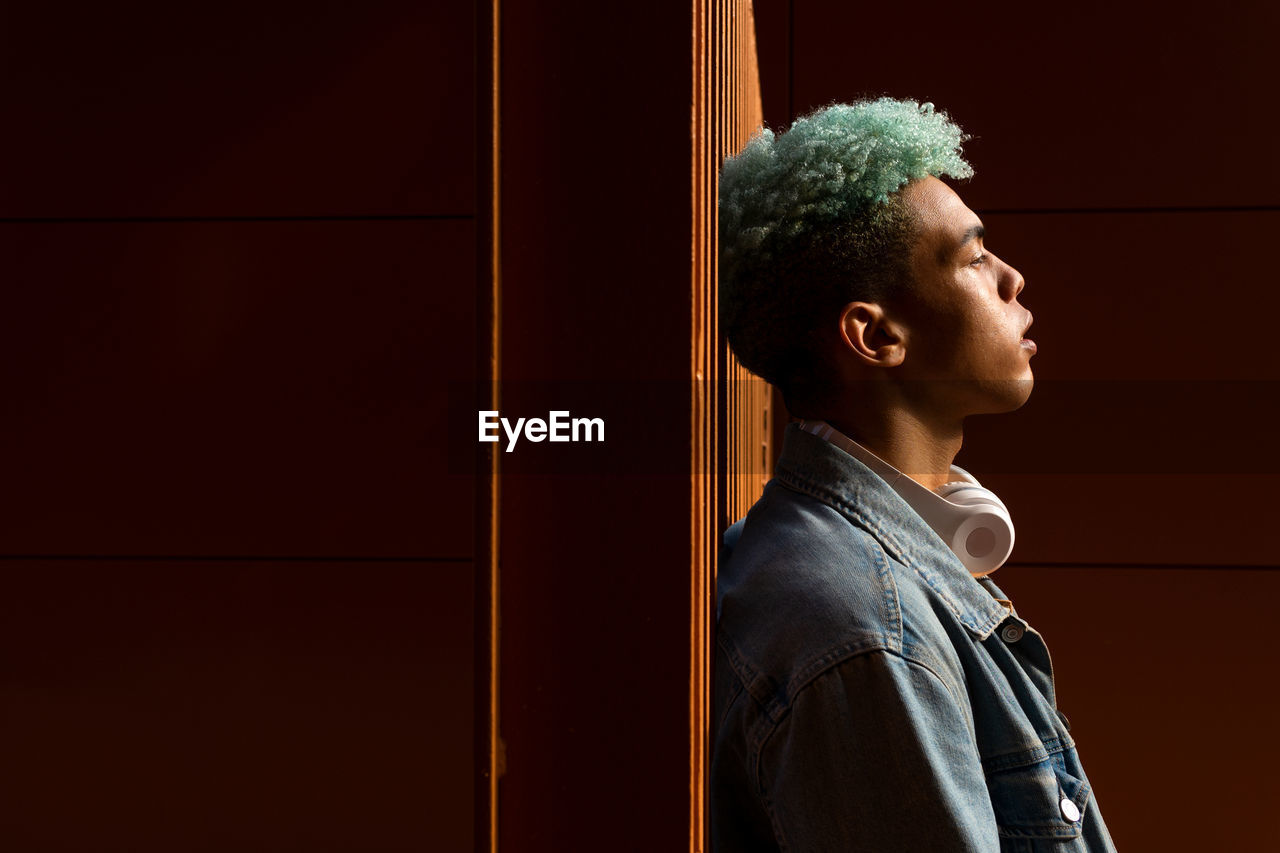 Side view of african american male hipster with blue curly hair leaning on wall in city looking away